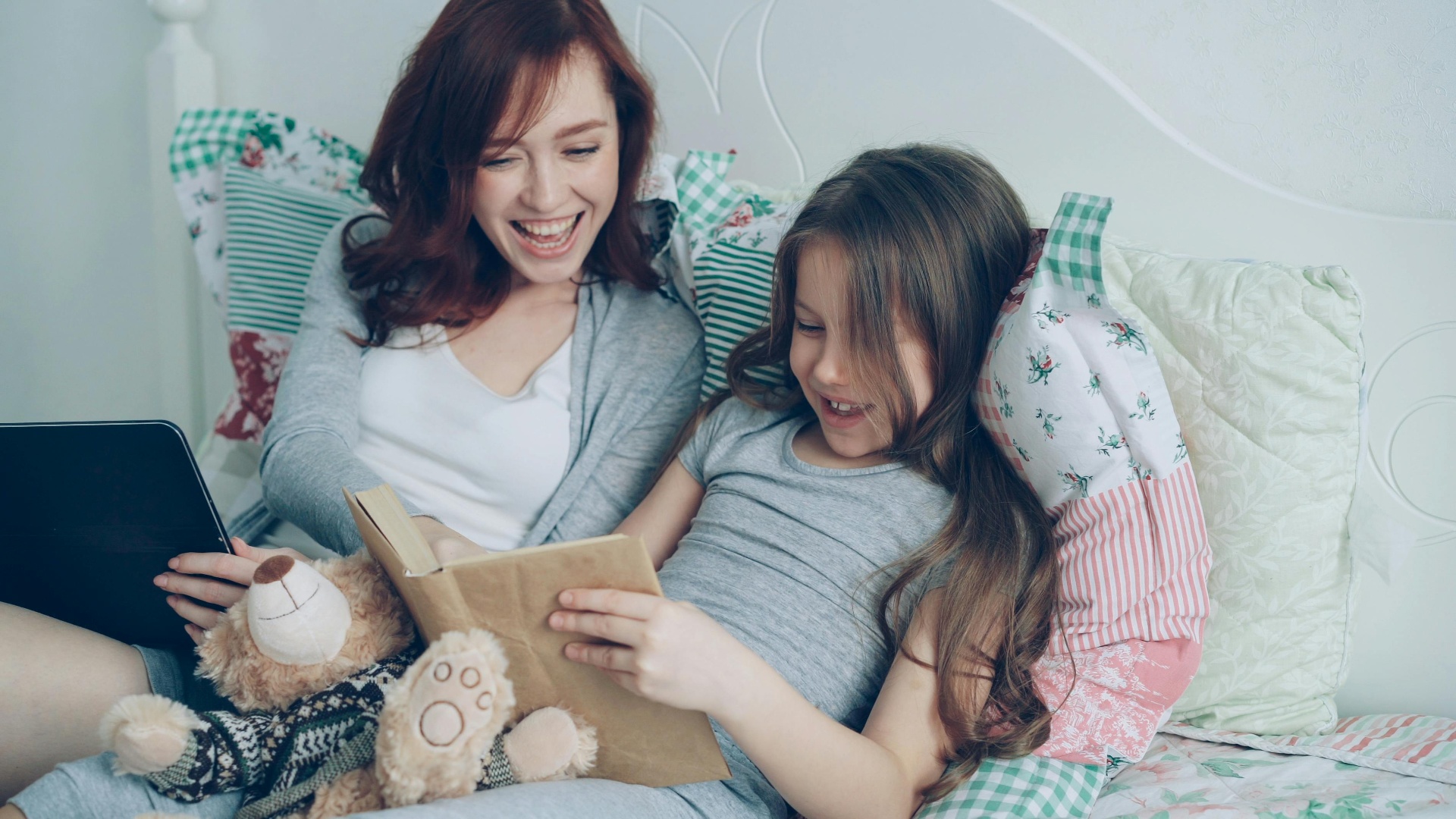 A mother and daughter reading together on a bed in soft, warm light.